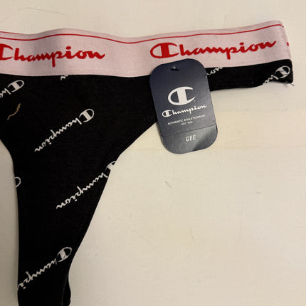 A pair of Champion women's thong underwear in black with a white script logo print, featuring a light pink waistband with red Champion branding and a dark gray brand tag.