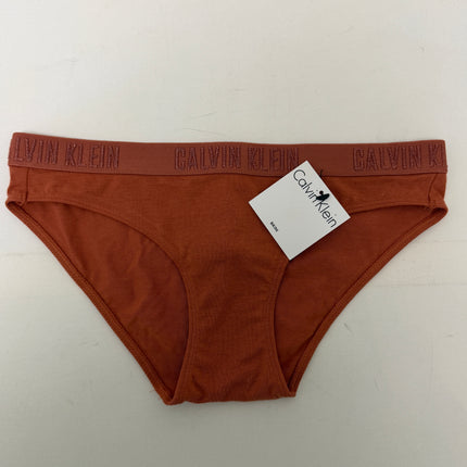 A pair of Calvin Klein ribbed briefs in a rust-orange color, featuring a matching elastic waistband with the brand name 'CALVIN KLEIN' repeated in tonal embroidery. A white paper brand tag is attached near the right hip, and the underwear is laid flat on a light off-white background.